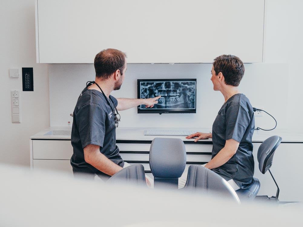 Dr. Hari Petsos und Dr. Carolin Petsos sitzen vor einem Bildschirm und besprechen das Röntgenbild eines Patienten oder einer Patientin.