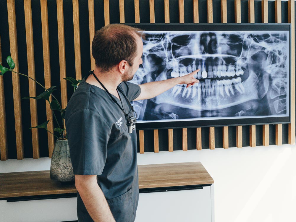 Zahnarzt Dr. Hari Petsos steht in seiner Praxis vor einem großen Bildschirm, auf dem ein Röntgenbild eines Gebisses angezeigt wird, und zeigt mit dem Zeigefinger auf einen Schneidezahn