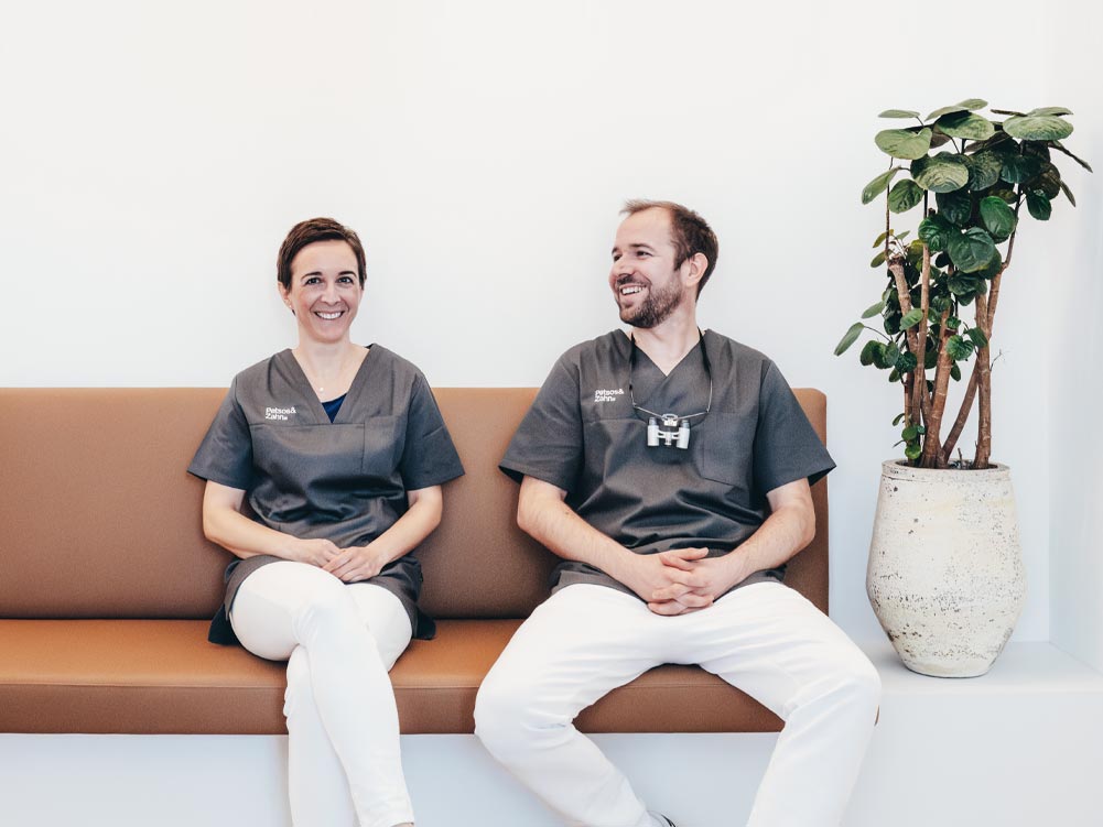 Auf einem hellbraunen Ledersofa sitzen Zahnärztin Dr. Carolin Petsos und Zahnarzt Dr. Hari Petsos fröhlich lächelnd in ihrer Zahnarztpraxis in Soest nebeneinander.