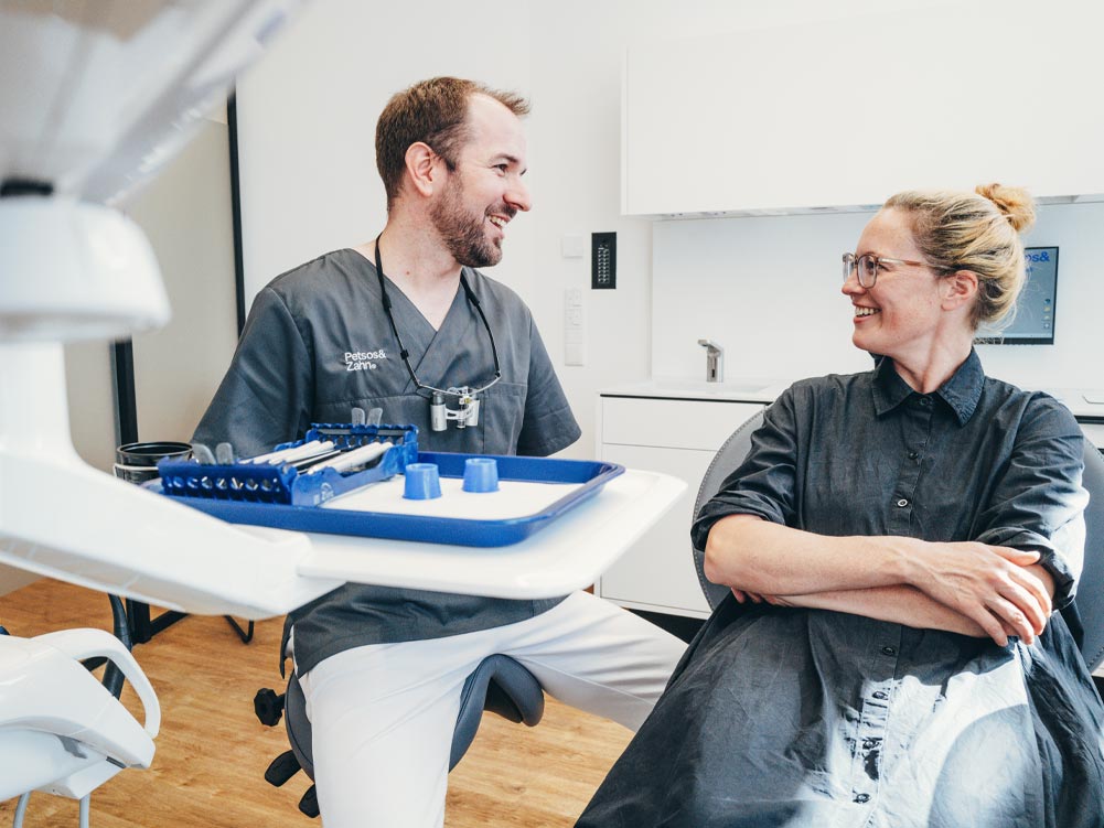 Zahnarzt Dr. Hari Petsos spricht lachend mit einer Patientin im Behandlungsstuhl. Beide sehen entspannt und zufrieden aus.