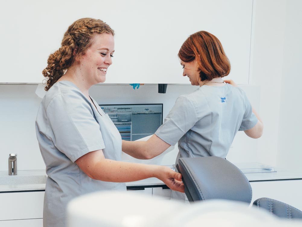 Zwei Mitarbeiterinnen bereiten fröhlich den Behandlungsplatz in der Praxis Petsos&Zahn vor. Sie lachen beide und haben offensichtlich Spaß.