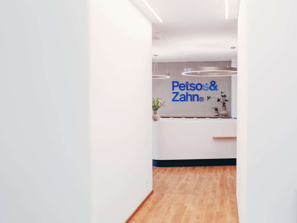 Man sieht den Empfangstresen der Praxis Petsos&Zahn. An der Wand hängt das Logo in blauen Acrylbuchstaben an der Wand. Der Boden ist aus hellem Holz, die Einrichtung hell und modern in weiß, grau und mit Pflanzen.